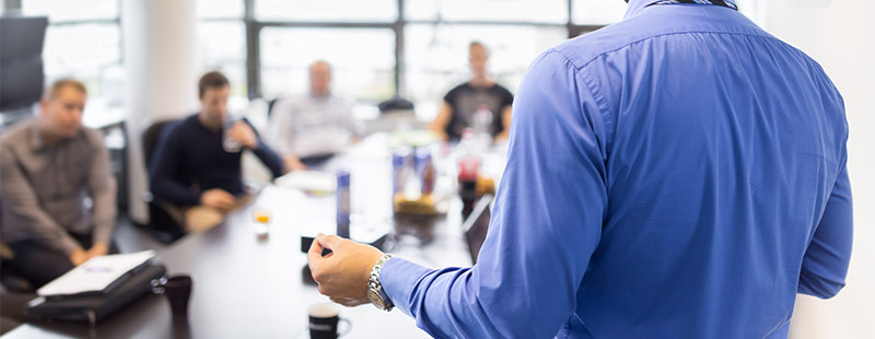 Selbstständige Ein Bild auf dem ein Mann in einem blauen Hemd vielen Menschen die an einem tisch sitzen im Hintergrund etwas erklärt