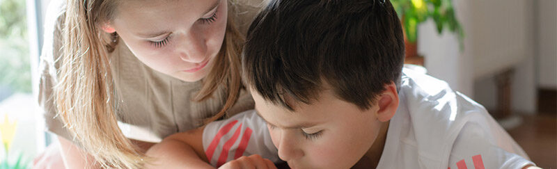 Ein Bild auf dem zwei Kinder in eine Zeitschrift schauen und das eine Kind mit dem Finger als Hilfe versucht vorzulesen