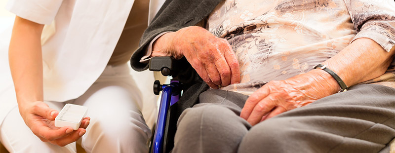 Ein Bild in dem eine Person im Rollstuhl sitzt und neben dran eine Pflegekraft eine Tabletten Schatulle hält