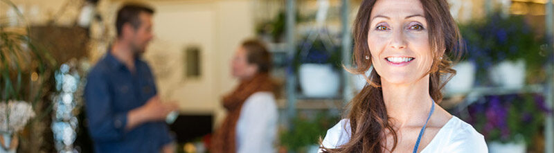 Eine lachende Frau mit einer Blauen Schürze im Vordergrund. Im Hintergrund sind 2 Personen die sich gerade unterhalten