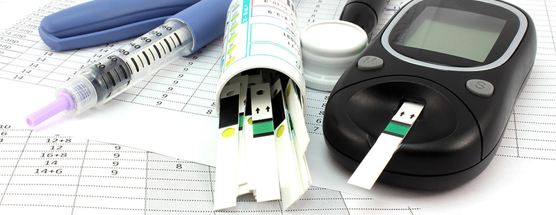 Ein Bild auf dem auf einem Tisch ein Diabetes Messgerät + die dazugehörigen Teststreifen und Unterlagen zum ausfüllen liegen