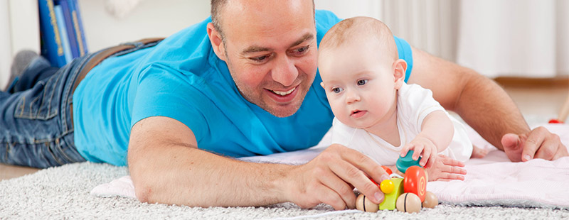 ein Bild auf dem ein Vater mit seinem Neugeborenen auf einem Teppich auf dem Boden liegt und spielt