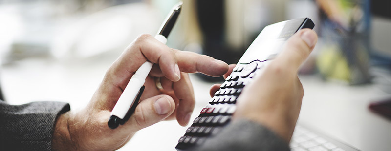 Ein Bild auf dem eine Hand den Taschenrechner hebt und in der anderen Hand einen Kugelschreiber hält, im Hintergrund sieht man eine Tastatur