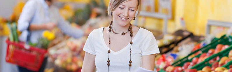 Eine Frau die ein Zettel in der Hand hält und Ihn anschaut, im Hintergrund befindet sich ein Markt mit viel Obst und Gemüse
