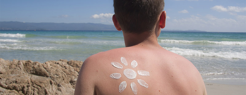 Ein Bild wo eine am Strand sitzt und auf dem Rücken mit Sonnencreme eine Sonne gemalt hat, im Vordergrund befindet sich noch das Meer