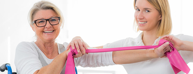Eine Frau im Rollstuhl und eine Frau daneben die mit der Frau im Rollstuhl Übungen mit dem Terraband macht