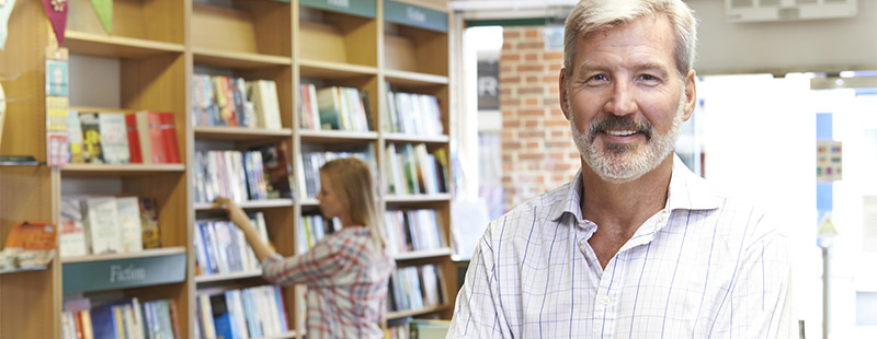 Ein lächelnder Mann im Vordergrund und im Hintergrund ist eine Frau die gerade an einem Bücherregal ist.