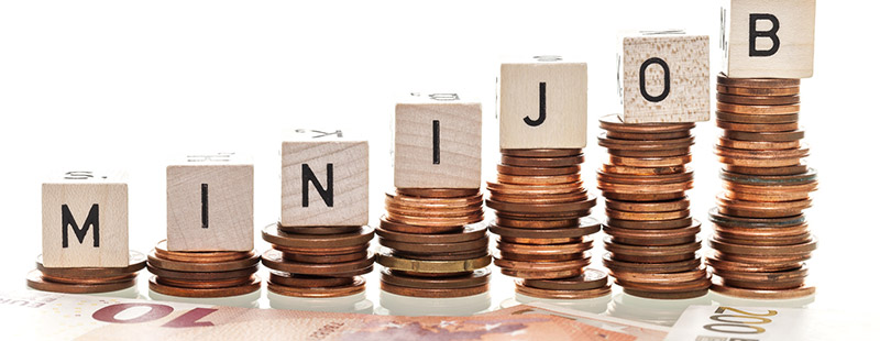 Ein Bild mit gestapeltem Kleingeld auf den Stapeln befinden sich Würfel mit jeweils den Buschstaben M-I-N-I-J-O-B unten drunter sieht man Geldscheine