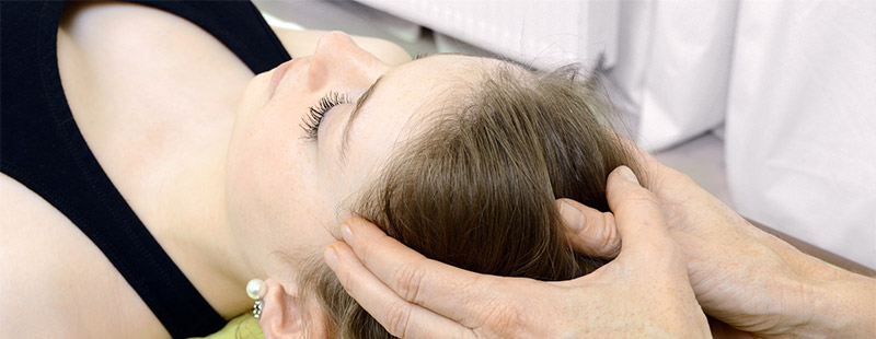 Ein Bild auf dem eine Frau auf einer Liege liegt und zwei Hände Ihren Kopf berühren