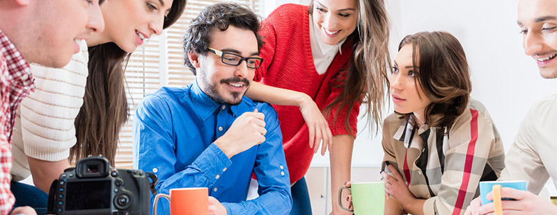 Mehrere Personen stehen oder sitzen eng zusammen und besprechen etwas ebenso ist eine Kamera und eine Orangene Tasse zu sehen