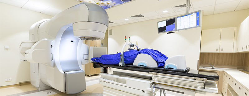 Auf diesem Bild sieht man ein großes Gerät in einem Krankenhaus Raum, ebenso sind Monitore und eine Krankenliege zu sehen.