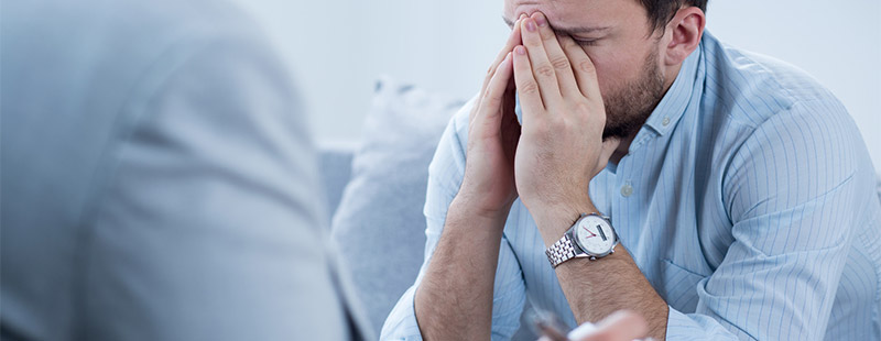 Ein Bild wo ein Mann auf einem Sofa sitzt und die Hände vor seinem Gesicht verschränkt. Eine weitere Person sitzt Ihm gegenüber.