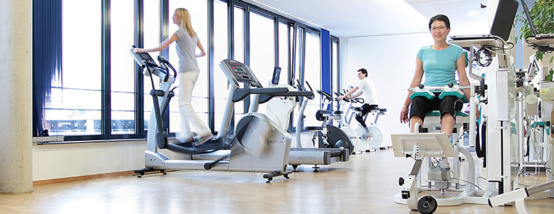 Mehrere Personen die auf verschiedenen Sport - Geräten sitzen und Sport machen. Im Hintergrund sieht man eine Fensterfront und eine Grüne Pflanze