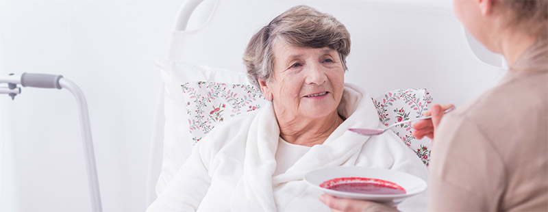 Eine ältere Dame die im Pflegebett sitzt und gerade von einer Pflegekraft das essen bekommt