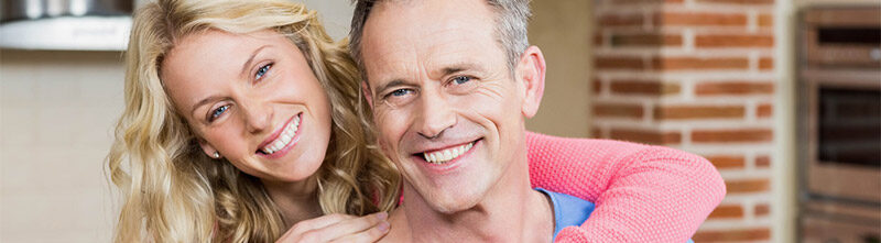 Ein Bild auf dem ein Mann lacht und eine Frau die hinter seinem Rücken sich nach vorne beugt und ebenso lacht. Im Hintergrund sieht man eine Mauer und Küchengeräte.