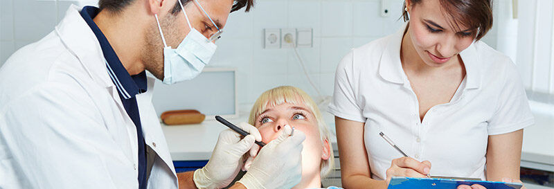 Eine Frau die auf einem Zahnarztstuhl liegt, links von Ihr ist ein Zahnarzt der in Ihren Mund schaut, rechts von Ihr ist eine Helferin die auf einem Zettel mit Ihrem Kugelschreiber alles notiert