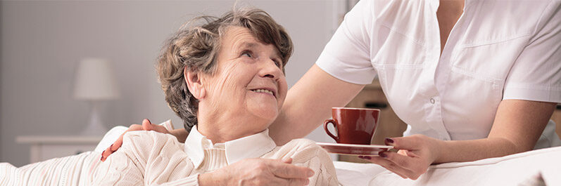 Eine Ältere Dame die auf dem Sofa sitzt und gerade von einer Pflegekraft einen Espresso gereicht bekommt