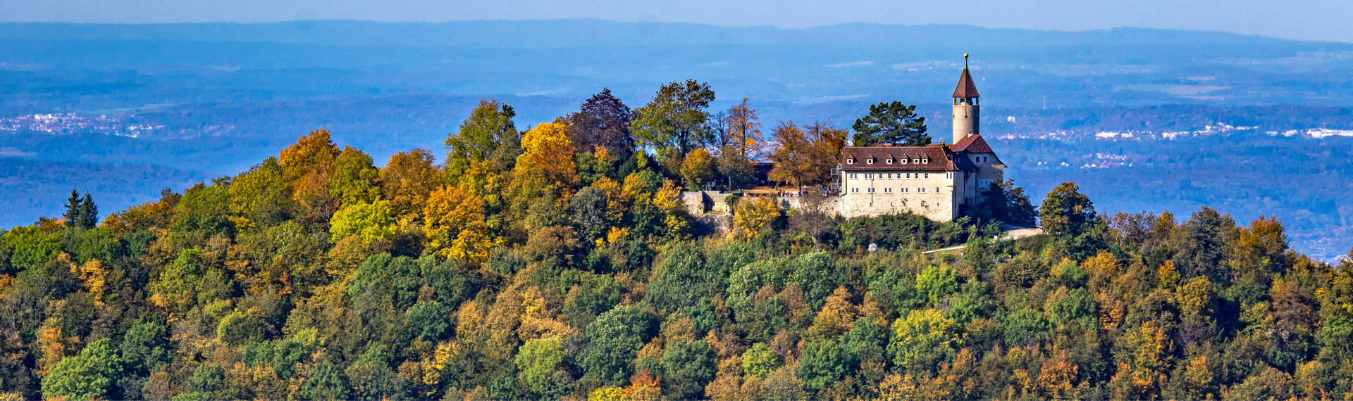 ein freundliches Bild auf dem die Burg Teck zu sehen ist, sie ist von bunten Bäumen umgeben. In der ferne sieht man die umliegende Dörfer.