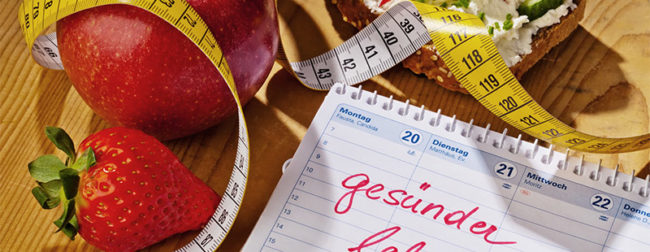 Ein Bild mit einem Holztische auf dem Obst liegt mit einem Maßband und einem Kalender auf dem Kalender steht gesünder leben