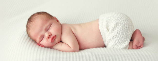Ein Bild in dem ein Baby auf einem Weißen Tuch liegt. Das Baby hat nur eine weiße Hose an und liegt mit dem Kopf auf seinen kleinen Händen